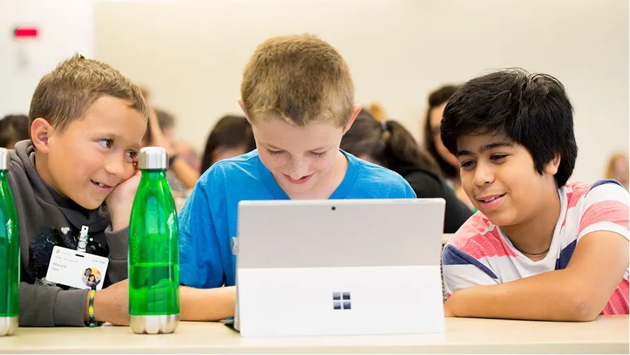 Drei Kinder sitzen an einem Tisch und schauen auf einen Laptop. Grüne Wasserflaschen stehen neben ihnen.