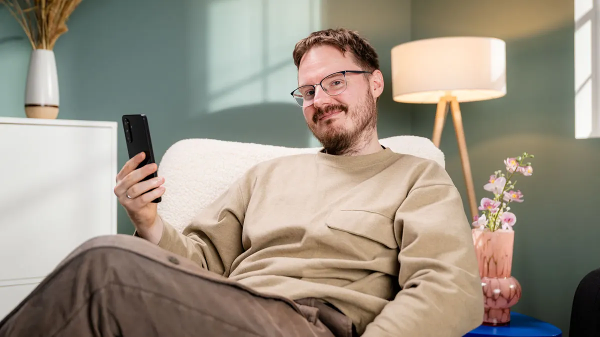 Michael sitzt mit dem Samsung Galaxy A26 5G in der Hand entspannt in einem Wohnzimmer und lächelt.