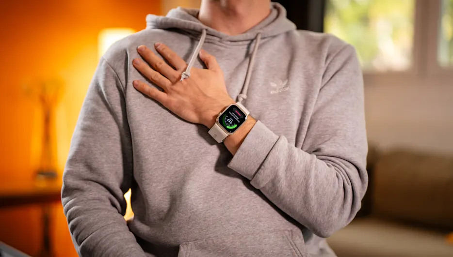 Mann mit grauer Kapuzenjacke und Smartwatch am Handgelenk, Hand liegt auf der Brust, heller Hintergrund.