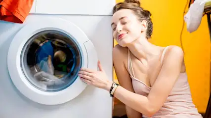 Eine Frau in einem Top lehnt an einer Waschmaschine mit Wäsche im Inneren vor einer gelben Wand.