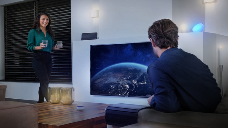 Eine Frau mit Getränken steht; ein Mann sitzt vor einem Fernseher, der einen blauen Planeten zeigt. Eine blaue Lampe leuchtet.