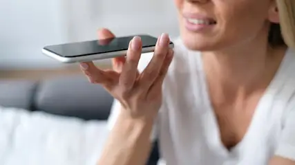 Eine Person hält ein schwarzes Smartphone in der Hand, während sie spricht. Sie trägt ein weißes Oberteil.