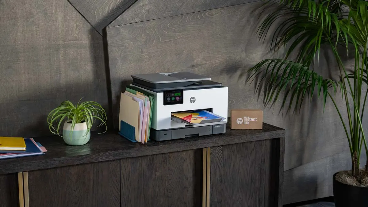 Ein Drucker steht auf einem dunklen Holzschrank, daneben Akten, eine Pflanze und eine Instant-Ink-Box. Im Hintergrund eine Wand und eine Palme.