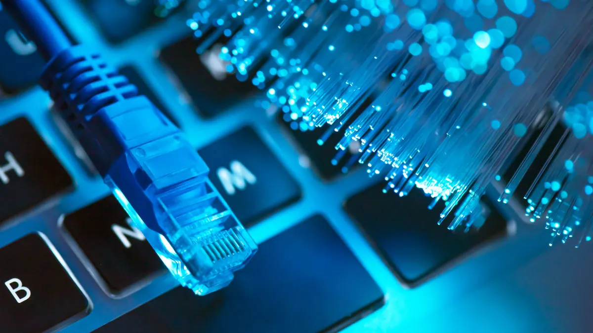 Der Stecker eines LAN-Kabels und Glasfasern liegen auf einer blau beleuchteten Laptop-Tastatur.