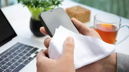 Ein schwarzes Handy wird mit einem weißen Tuch geputzt. Im Hintergrund sind Laptop, Tee, Kuchen und eine Pflanze zu sehen.