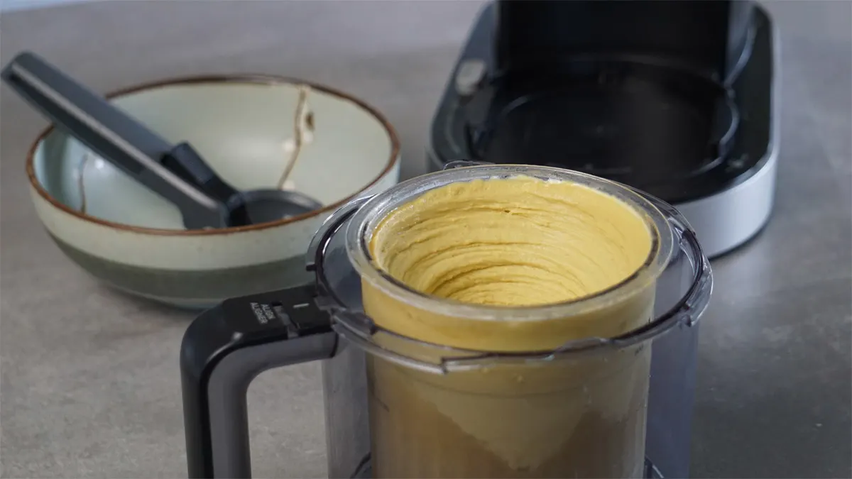 Ein cremiges Mangosorbet im Behälter der Ninja Creami Deluxe, im Hintergrund stehen die Eismaschine und eine Schüssel.