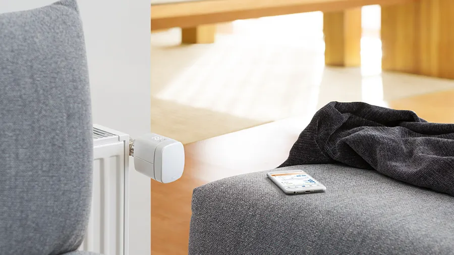 Ein graues Sofa mit einer dunklen Decke und einem Smartphone darauf. Im Hintergrund ein Heizkörper mit Thermostat.