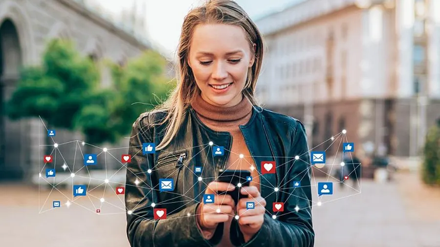 Eine Frau mit Jacke benutzt ein Smartphone, umgeben von Social-Media-Symbolen vor einem Gebäude.