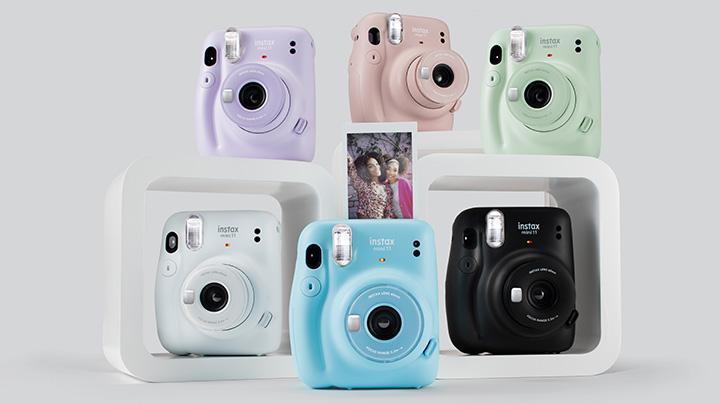 Sieben Sofortbildkameras in Lila, Rosa, Grün, Weiß, Blau und Schwarz stehen in weißen Regalen mit einem Foto auf der blauen Kamera.