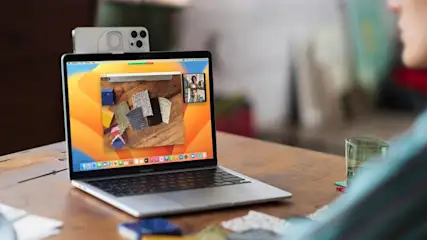 Ein Laptop auf einem Holztisch zeigt eine Videoanruf-App. Ein Smartphone ist an der Oberseite des Laptop-Bildschirms befestigt. Ein Glas ist rechts.