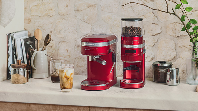 Auf einer Theke stehen eine rote Kaffeemühle, eine Espressomaschine, ein Glas Kaffee und Küchenutensilien.