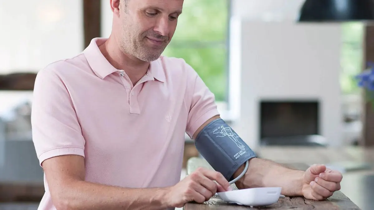 Ein Mann sitzt an einem Holztisch und misst gerade seinen Blutdruck.