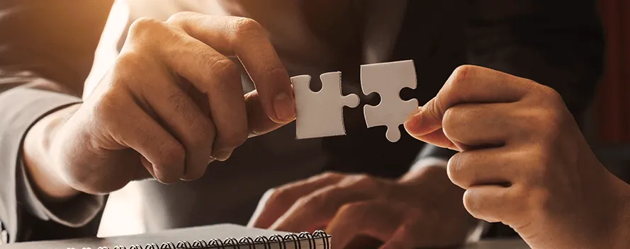 Zwei Hände halten weiße Puzzleteile, die kurz davor stehen, zusammengefügt zu werden. Im Vordergrund liegt ein Notizbuch.