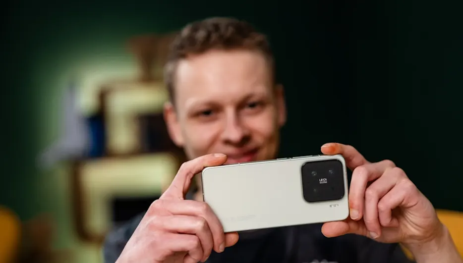 Ein Mann hält ein helles Smartphone mit einer schwarzen Kamera in horizontaler Ausrichtung vor sich. Der Hintergrund ist unscharf.