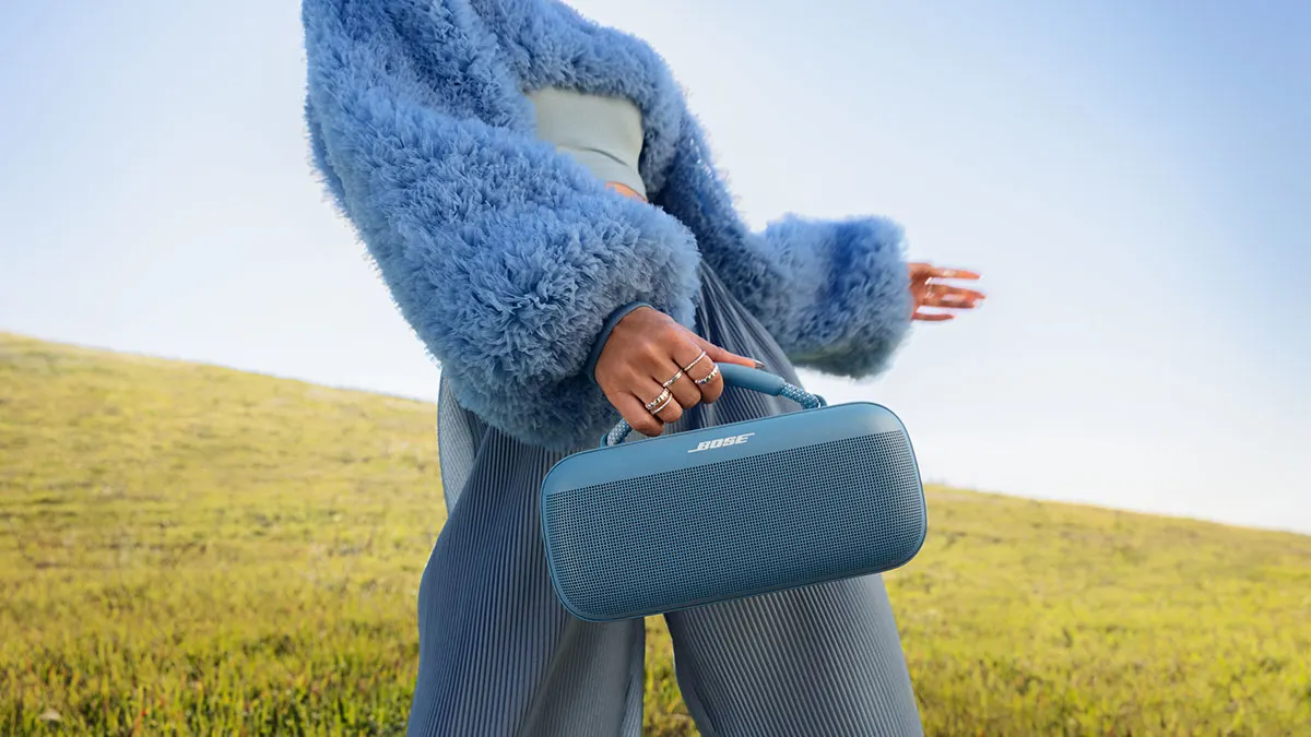 Eine Person hält einen blauen Bose-Lautsprecher vor einer Wiese unter blauem Himmel. Sie trägt einen blauen Pelzmantel und eine plissierte Hose.