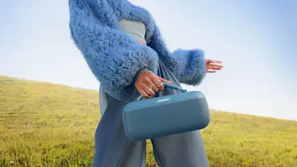 Eine Person hält einen blauen Bose-Lautsprecher vor einer Wiese unter blauem Himmel. Sie trägt einen blauen Pelzmantel und eine plissierte Hose.