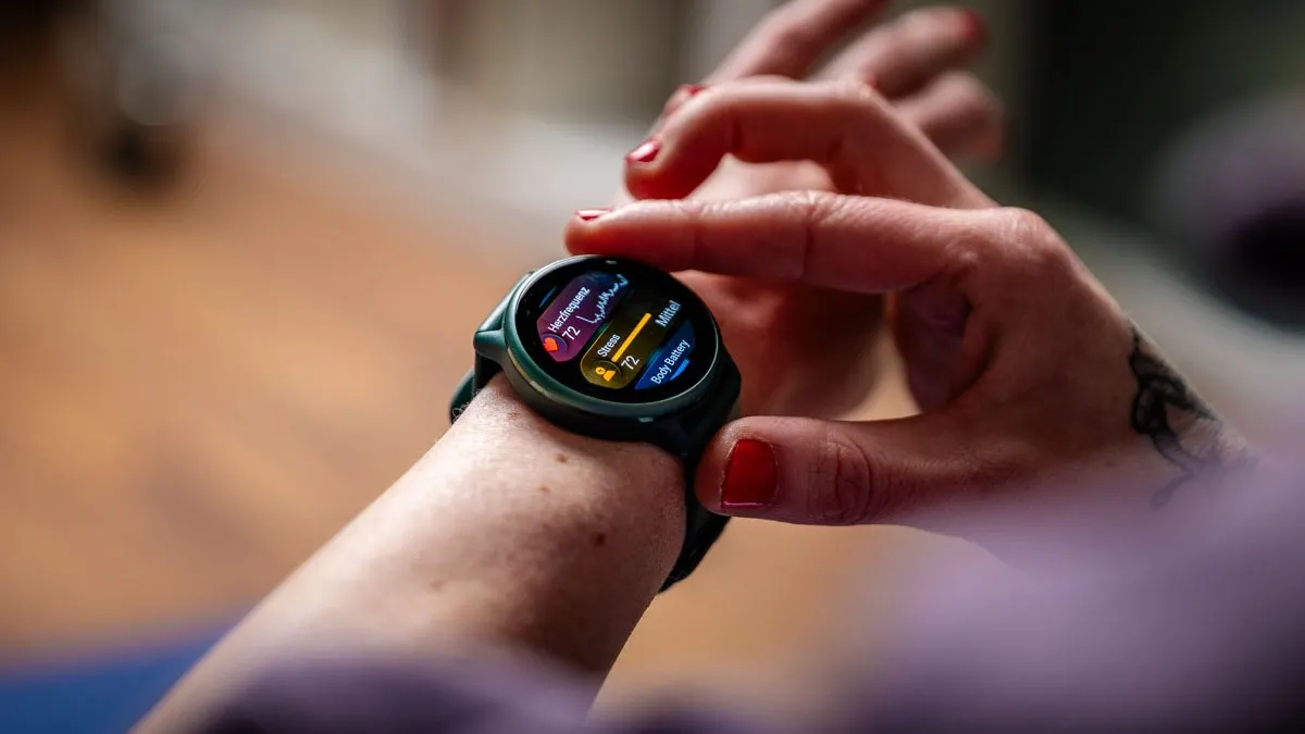 Eine Person mit rotem Nagellack berührt eine grüne Smartwatch am Handgelenk. Die Uhr zeigt Gesundheitsdaten.