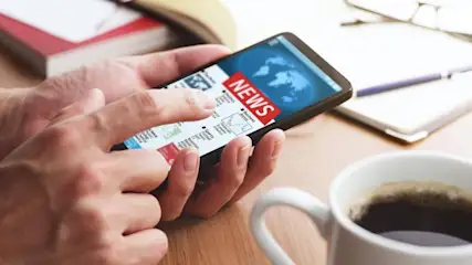 Eine Person hält ein Smartphone mit einer Nachrichten-App in der Hand, daneben eine Kaffeetasse, Notizbuch und Stifte.