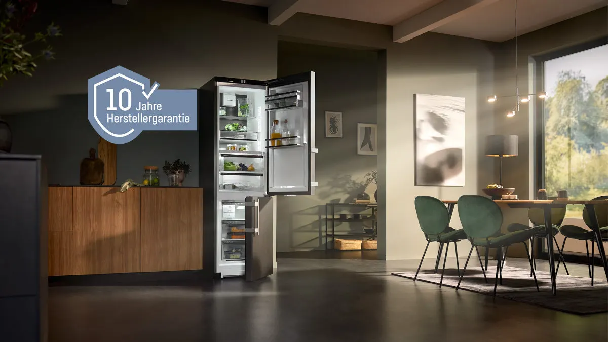 Ein Kühlschrank mit offener Tür steht in einem Raum mit Esstisch und Holzschrank. Ein Logo für 10 Jahre Garantie ist sichtbar.