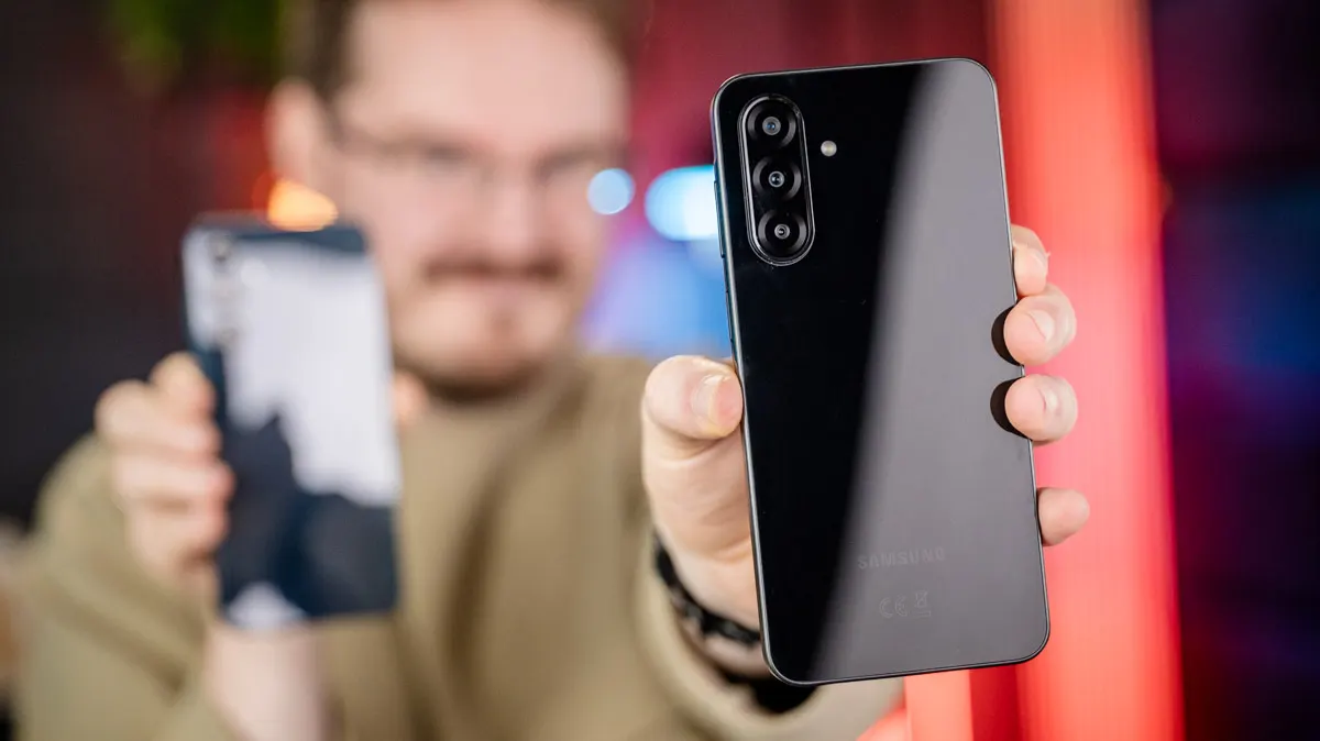 Michael hält das Samsung Galaxy A26 in der linken Hand und streckt seinen Arm nach vorn. In seiner anderen Hand hält er das Samsung Galaxy A16, das im Hintergrund nur verschwommen zu sehen ist.