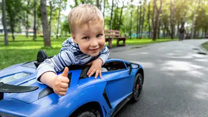 Ein Kind im gestreiften Hemd sitzt in einem blauen Spielzeugauto auf einem grauen Weg. Bäume und eine Bank sind im Hintergrund.