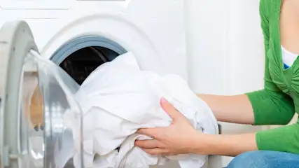 Eine Person in grüner Kleidung räumt weiße Wäsche in eine weiße Waschmaschine ein. Die Tür ist offen.