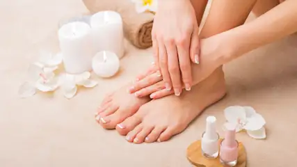 Hände und Füße mit weiß lackierten Nägeln. Im Hintergrund: Kerzen, Handtuch, Nagellack und weiße Orchideenblüten.