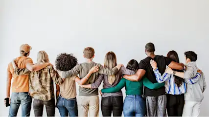 Eine Gruppe von neun Personen steht mit verschränkten Armen vor einer weißen Wand. Sie tragen Freizeitkleidung und blicken nach vorne.