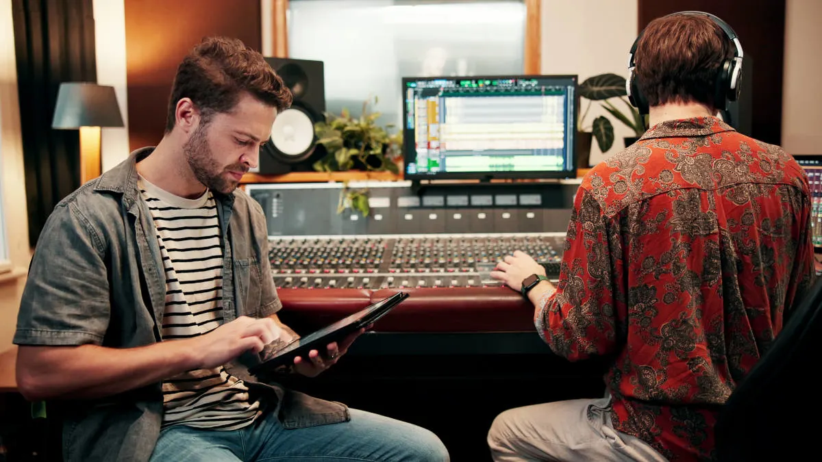 Zwei Personen sitzen vor einem Mischpult. Eine schaut auf ein Tablet, die andere trägt Kopfhörer und arbeitet am Mischpult.
