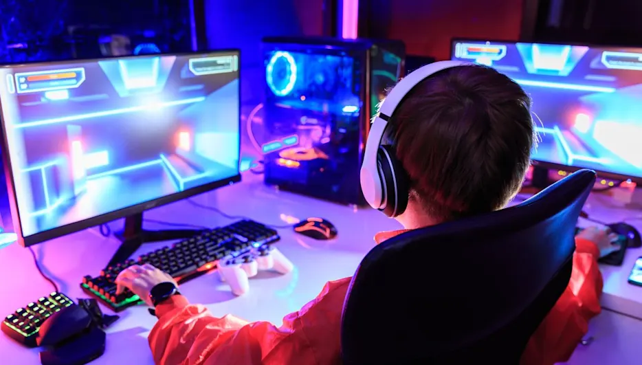 Ein Junge mit Kopfhörern sitzt vor zwei Monitoren und spielt ein Videospiel mit Tastatur und Maus. Ein Computer steht auch auf dem Tisch.