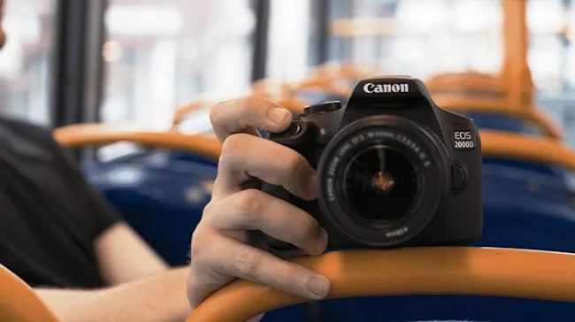 CANON EOS 2000D Kit Spiegelreflexkamera, 24,1 Megapixel, 18-55 mm Objektiv (EF-S, IS II), WLAN, Schwarz Eine Canon EOS 2000D Kamera wird von einer Hand auf einer orangefarbenen Stange gehalten. Blaue und weiße Elemente im Hintergrund.