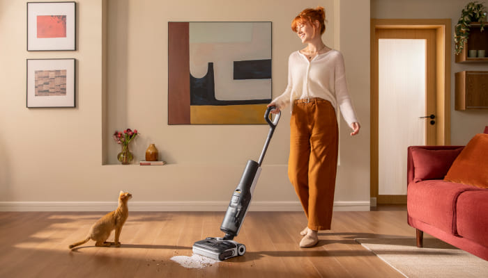 Eine Frau benutzt einen Staubsauger auf einem Holzboden, während eine Katze zusieht. Ein rotes Sofa steht im Hintergrund.