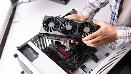 Eine Person hält eine schwarze Grafikkarte mit drei Lüftern über ein offenes, weißes Computergehäuse.