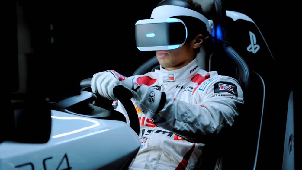 Ein Gamer in einem Rennanzug sitzt mit PlayStation-VR-Headset in einem Rennsitz und spielt ein Rennspiel. 