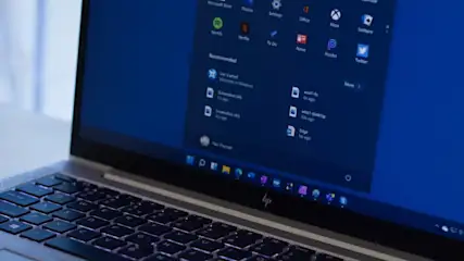 Ein Laptop zeigt ein blaues Windows-Startmenü mit App-Symbolen und Dateinamen auf einem silbernen Gehäuse mit schwarzer Tastatur.
