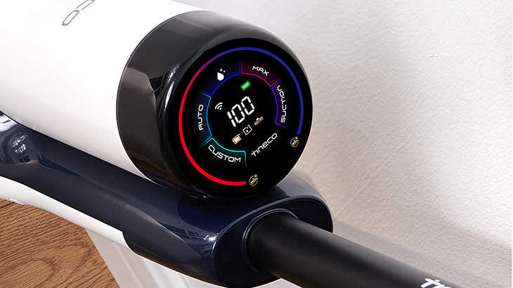 Ein kabelloser Staubsauger mit rundem Display zeigt 99% Akku auf hellem Untergrund mit Flecken. "AUTO" Modus ist aktiviert.