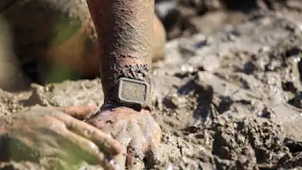Eine Hand und ein Unterarm mit einer eckigen Uhr sind mit Schlamm bedeckt, im Hintergrund ist mehr Schlamm.