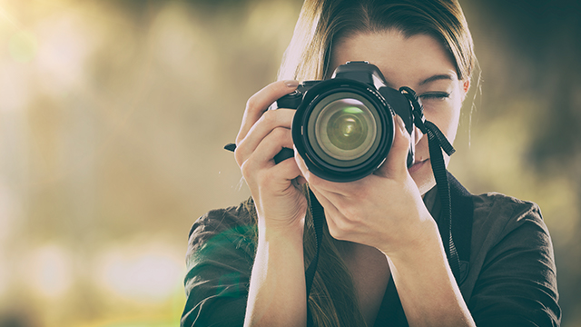 Eine Frau hält eine schwarze Kamera mit Gurt vor ihr Gesicht. Der Hintergrund ist unscharf und hell.