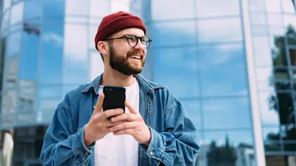 Ein Mann mit Brille und roter Mütze hält ein Smartphone vor einem Glasgebäude. Er trägt ein Jeanshemd über einem weißen T-Shirt.