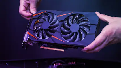 Eine Grafikkarte mit zwei Lüftern wird von zwei Händen über einem Computergehäuse gehalten. Orangefarbene Akzente sind sichtbar.