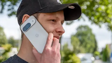 Ein Mann mit Kappe hält ein graues Smartphone mit blauer Kamera am Ohr, im Hintergrund Bäume.
