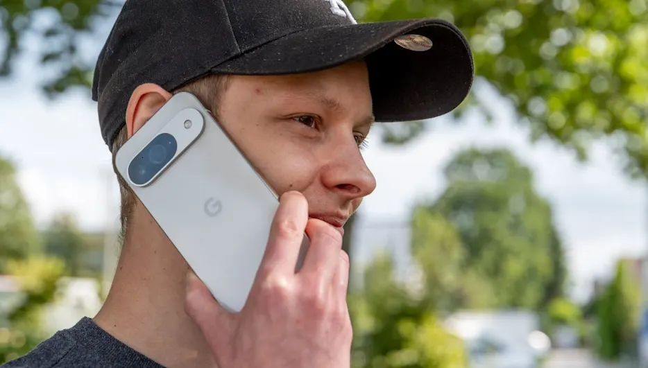 Ein Mann mit Kappe hält ein graues Smartphone mit blauer Kamera am Ohr, im Hintergrund Bäume.