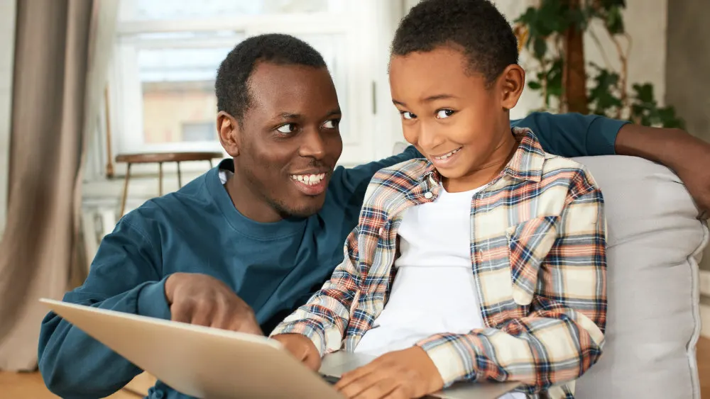 Ein Erwachsener und ein Kind sitzen zusammen auf einem Sofa und benutzen einen Laptop in einem hellen Raum.