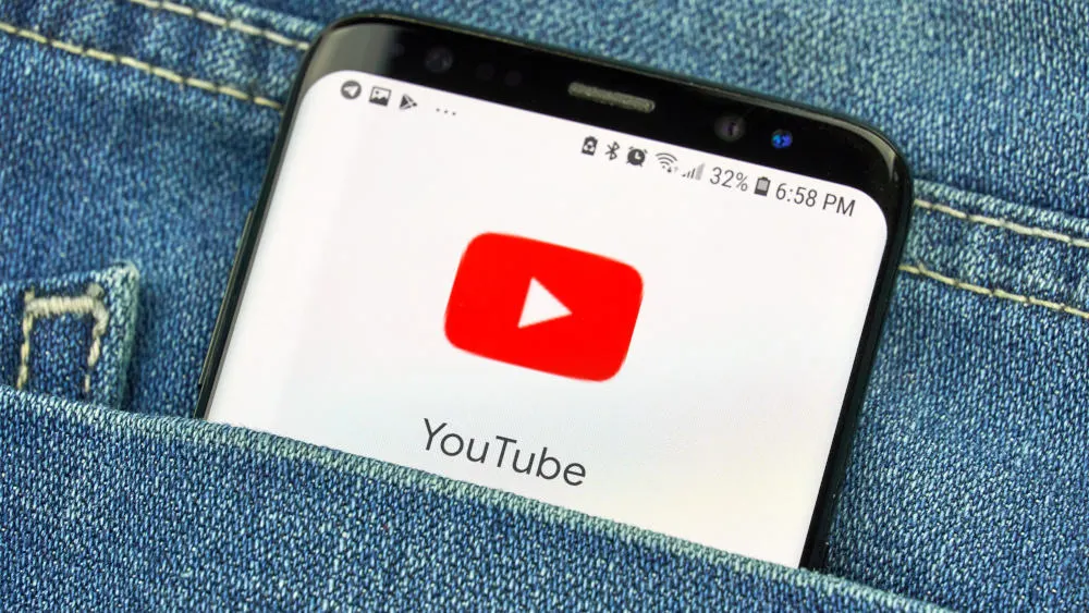 Ein Smartphone mit dem YouTube-Logo auf dem Bildschirm steckt in einer blauen Jeanshosentasche. Die Akkuladung beträgt 32%.