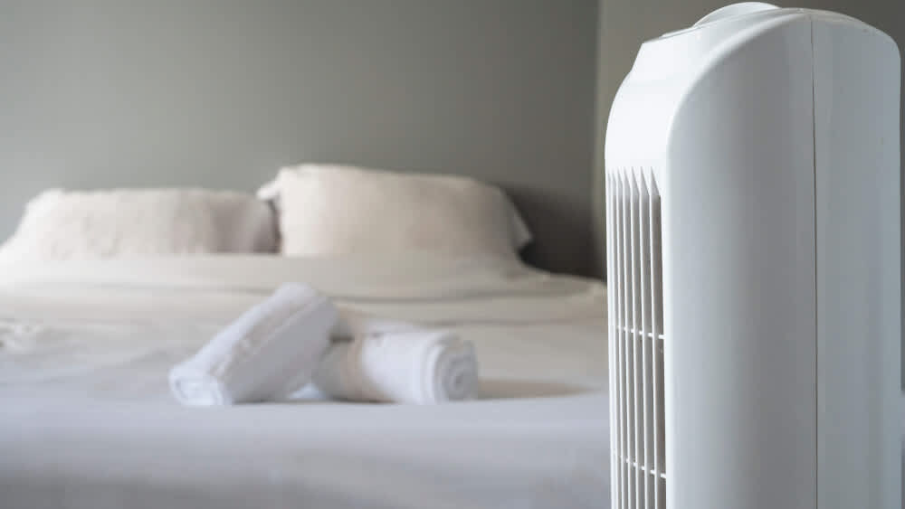 Ein Ventilator steht im Vordergrund und zwei Handtücher liegen auf einem Bett im Hintergrund.
