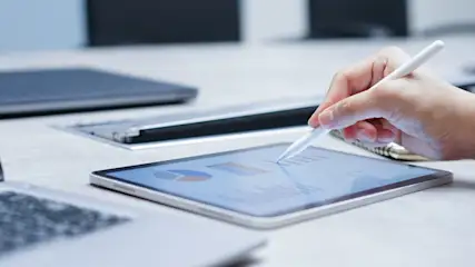 Eine Hand bedient ein Tablet mit einem weißen Stift. Das Tablet zeigt Diagramme. Im Hintergrund sind Laptops und ein Notizbuch sichtbar.