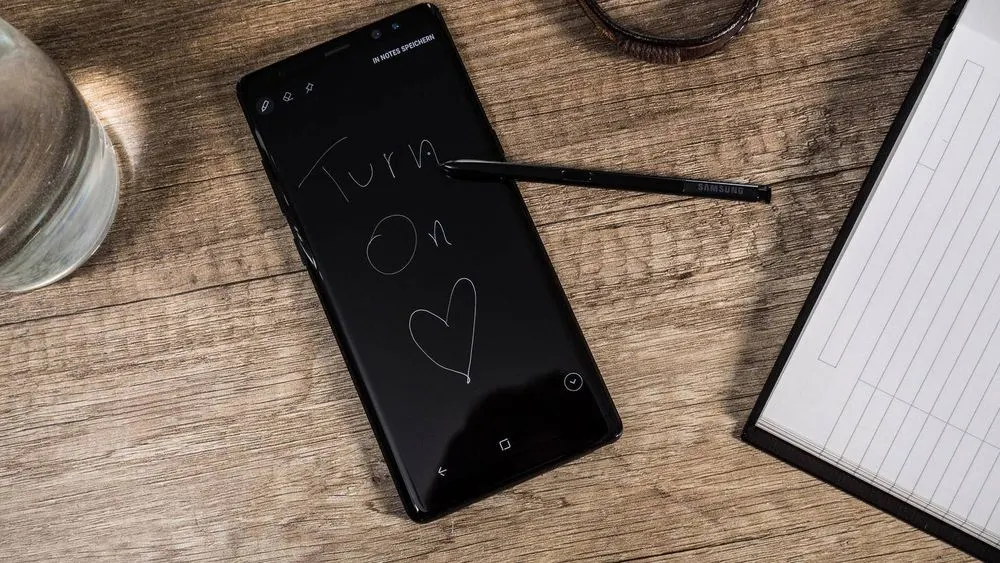 Ein schwarzes Smartphone mit der Aufschrift "Turn On" und einem Herz liegt auf einem Holztisch neben einem Stift, einem Notizbuch und einem Glas Wasser.