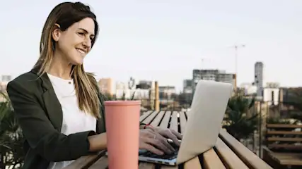 Frau tippt auf Laptop an Holztisch, rosa Becher daneben. Gebäude und Himmel im Hintergrund. Sie trägt einen grünen Blazer.