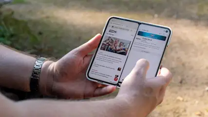 Eine Person hält ein faltbares Smartphone mit zwei Bildschirmen, auf denen Webseiten angezeigt werden. Eine silberne Uhr ist am Handgelenk sichtbar.