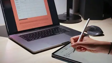 Ein Laptop zeigt eine Tabelle, daneben ein Tablet mit Stift in der Hand, eine Maus und ein schwarzer Lautsprecher im Hintergrund.